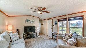 A living room with a ceiling fan, a television, and a large window. - Waterfront Home w/ Boat Slips in Northern Michigan (Gladwin)