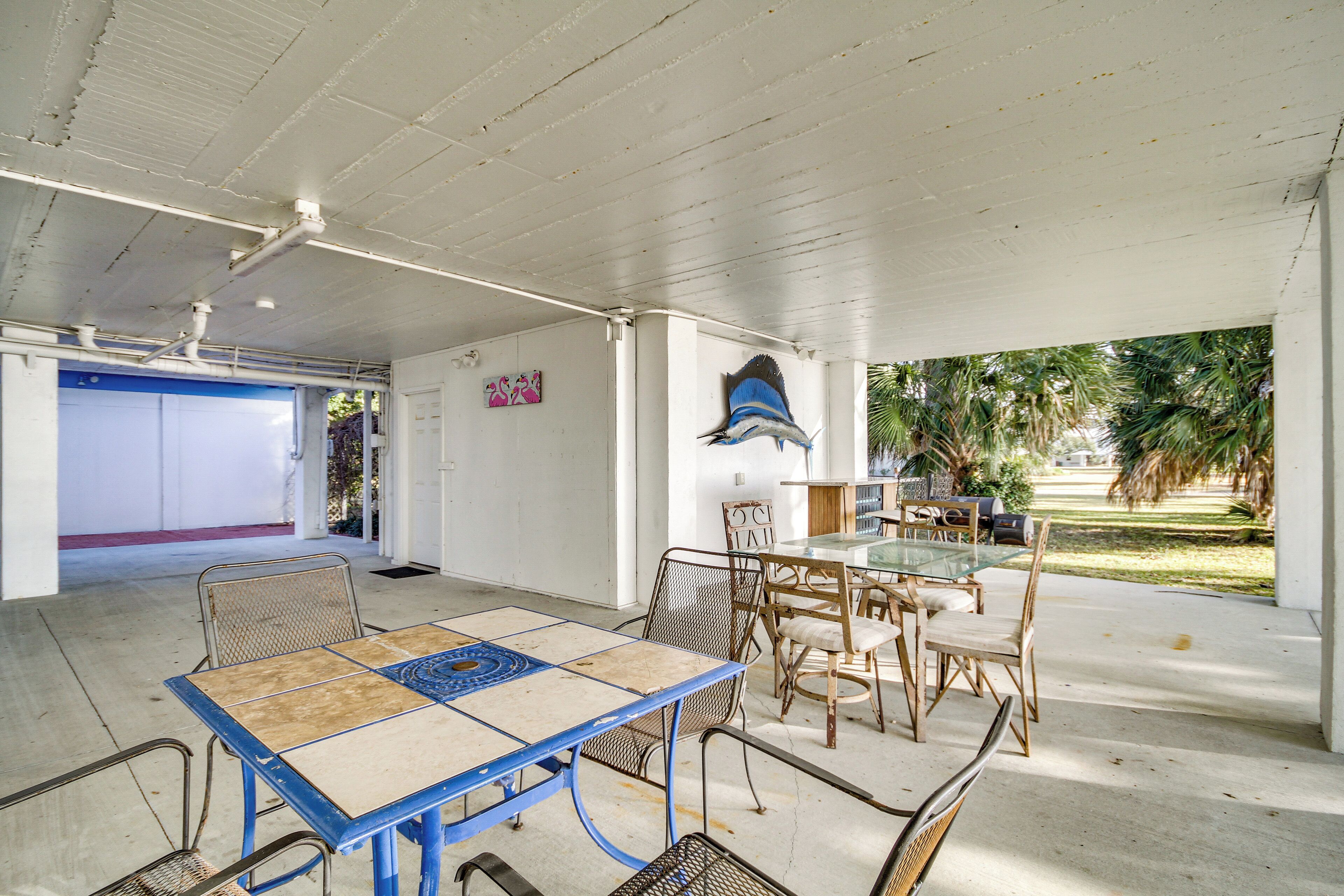 A covered outdoor dining area with a table and chairs, a ceiling with exposed beams, and a view of palm trees.
