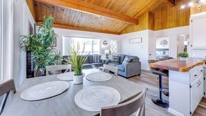 A spacious living area with a wooden ceiling, a grey sofa, a dining table with placemats, and a kitchen area with a bar counter. - Close to Skiing & Lake: Big Bear Mountain Getaway (Big Bear City)