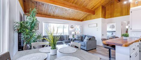 A spacious living area with a wooden ceiling, a grey sofa, a dining table with placemats, and a kitchen area with a bar counter.