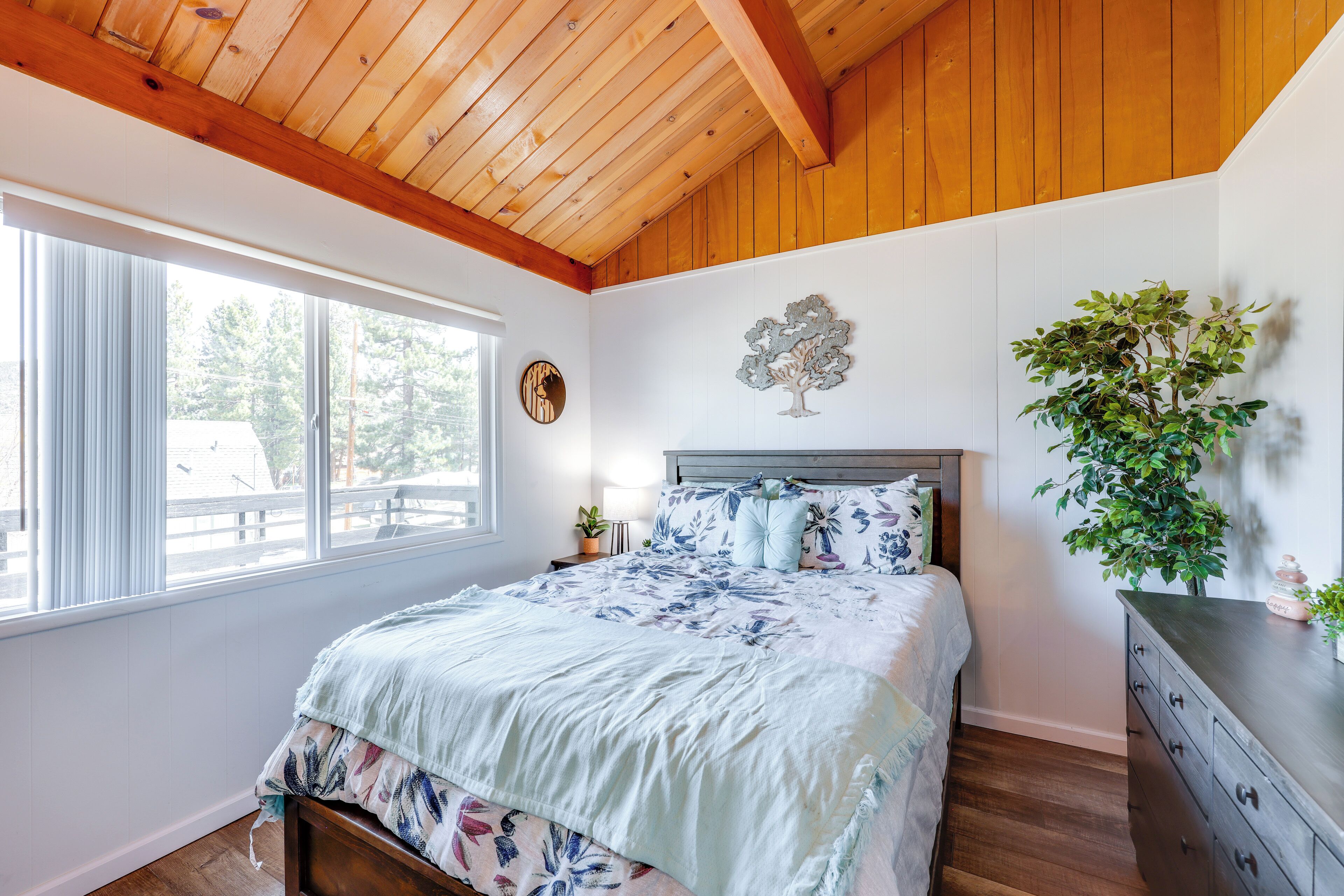 Un dormitorio con techo de madera, un ventanal grande, una cama con ropa de cama floral, una mesita de noche, un aparador y una planta en maceta.