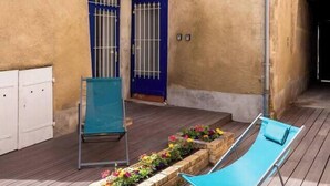 A wooden deck with a blue lounge chair and a white chair, a blue door, and a window with shutters. - Country apartment (Moulins)