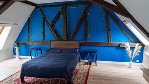 A bedroom with a sloped ceiling, a bed, two chairs, a small table, a potted plant, and a vase with dried pampas grass. - Country apartment (Moulins)