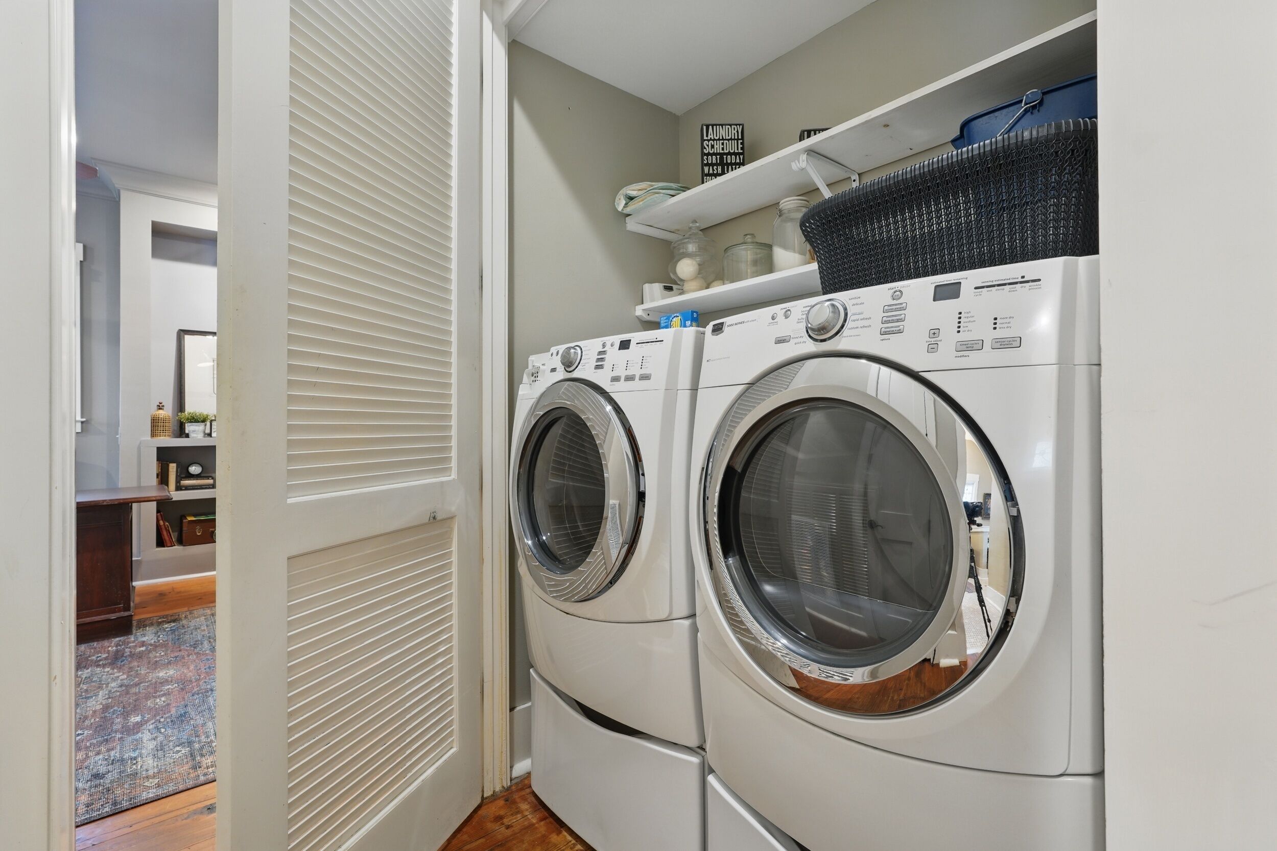 Un cuarto de lavandería con dos lavadoras de carga frontal apiladas, un estante con productos de limpieza y una puerta que da a otra habitación.