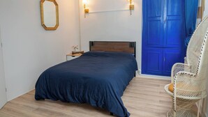 Quarto com cama, mesa de cabeceira, luminária na parede, porta azul e cadeira de vime.