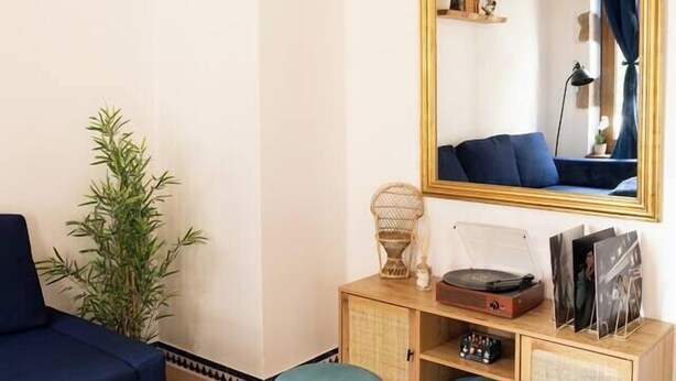Uma sala de estar com um sofá azul, uma mesa de centro branca, um banquinho azul-esverdeado, um armário de madeira com espelho e uma planta de bambu.