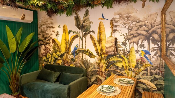 Una habitación con un sofá verde, una mesa de bambú y un mural de follaje tropical y aves.