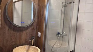 Un baño con tocador de madera, espejo redondo, lavamanos de piedra y ducha con mampara de vidrio y cabezal de ducha de bambú.