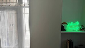 Una habitación con una ventana cubierta por cortinas traslúcidas, un letrero de neón en la pared y una silla en primer plano.