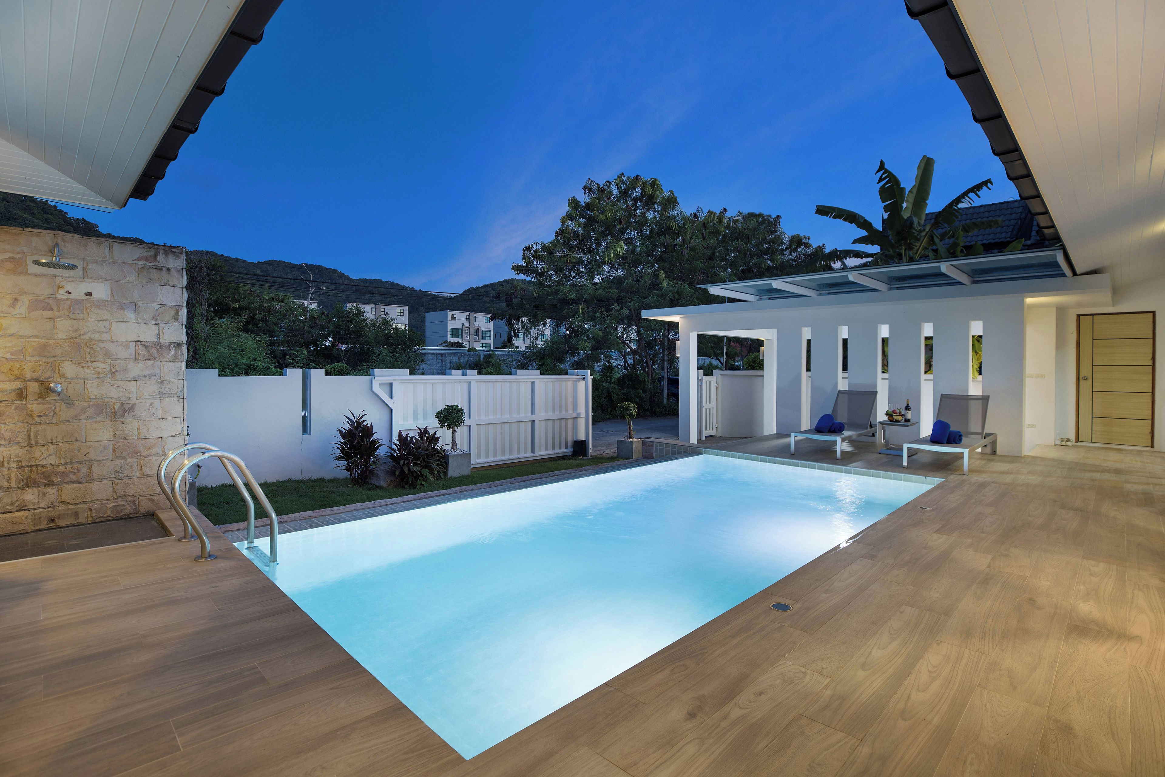 A modern indoor swimming pool with a clear view of the surrounding area, including a stone wall, a white fence, and a building with a balcony.