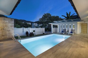 A modern indoor swimming pool with a clear view of the surrounding area, including a stone wall, a white fence, and a building with a balcony. - Designer 3BR Villa Silver Pool in Kamala (Kamala)