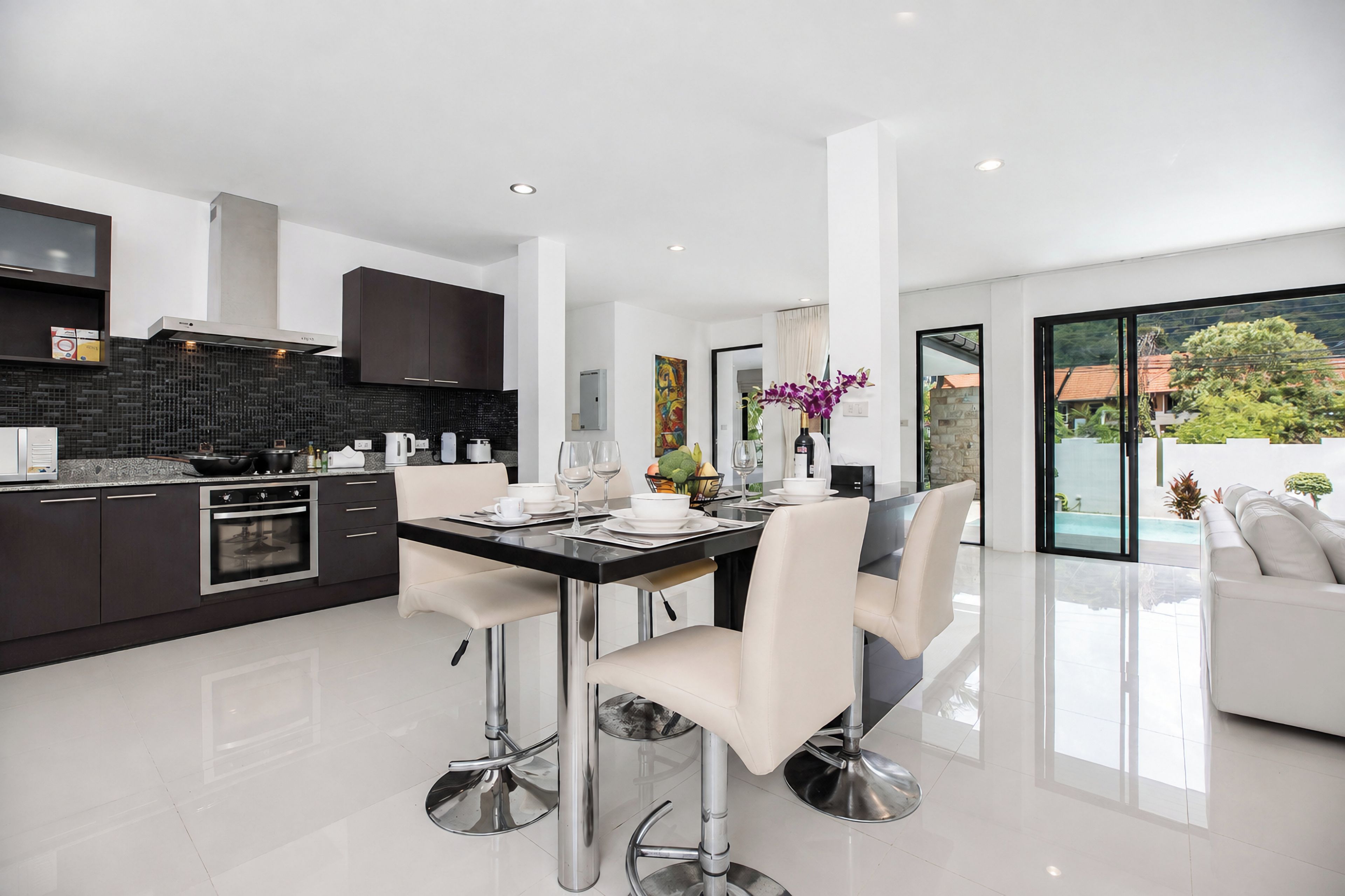 A modern kitchen with dark cabinetry, a stainless steel oven, and a dining area with a glass table and white chairs.