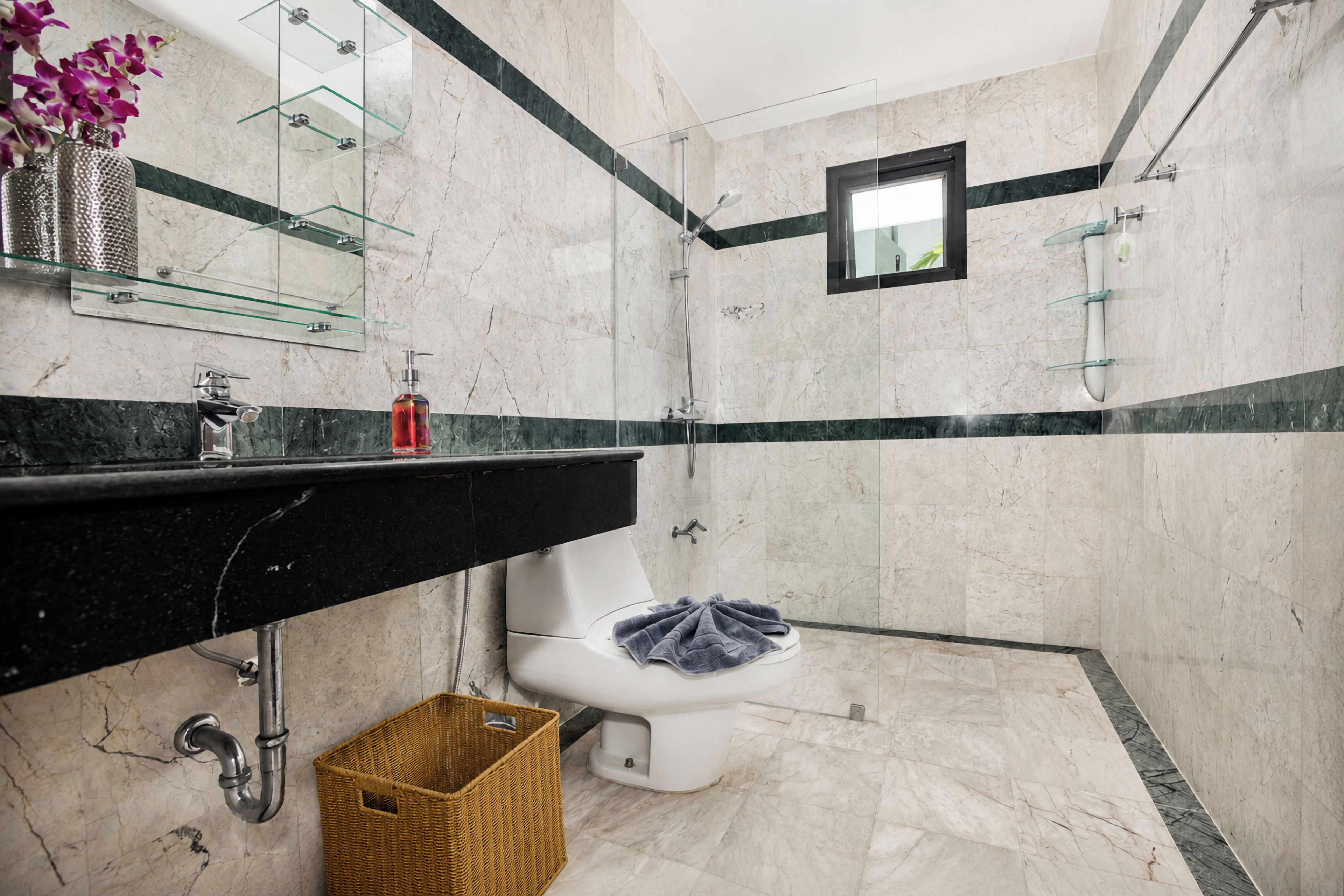 Egy modern fürőszoba márványburkolattal, fekete mosdópulttal, üveg zuhanykabinnal és egy törölközővel ellátott WC-vel.