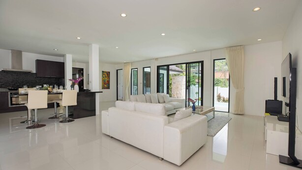 A modern living room with a white sofa, a dining area with a table and chairs, a kitchen with dark cabinets, and large windows with curtains. - Designer 3BR Villa Silver Pool in Kamala (Kamala)