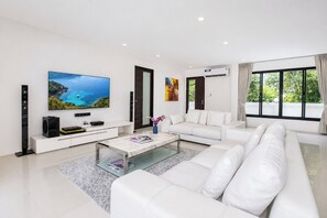 A modern living room with a white sofa, a coffee table, a flat-screen TV, and a large window with curtains. - Designer 3BR Villa Silver Pool in Kamala (Kamala)