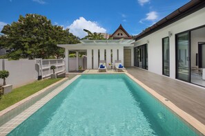 A rectangular swimming pool with a tiled edge, surrounded by a wooden deck. There are lounge chairs with blue cushions and a glass-enclosed patio area.