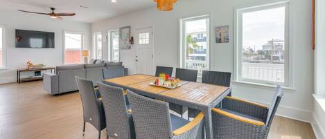 A spacious living area with a dining table, wicker chairs, a ceiling fan, and a large window offering a view of the city.