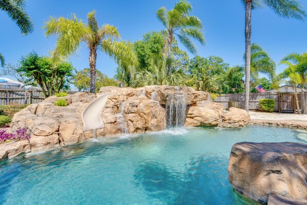 A clear pool with a waterfall feature, surrounded by palm trees and a wooden fence. - Private Outdoor Oasis w/ Pool: Home in Longwood! (Longwood)