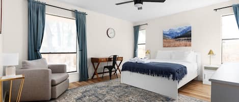 Een slaapkamer met een bed, een bureau, een stoel, een klok en een raam met gordijnen.