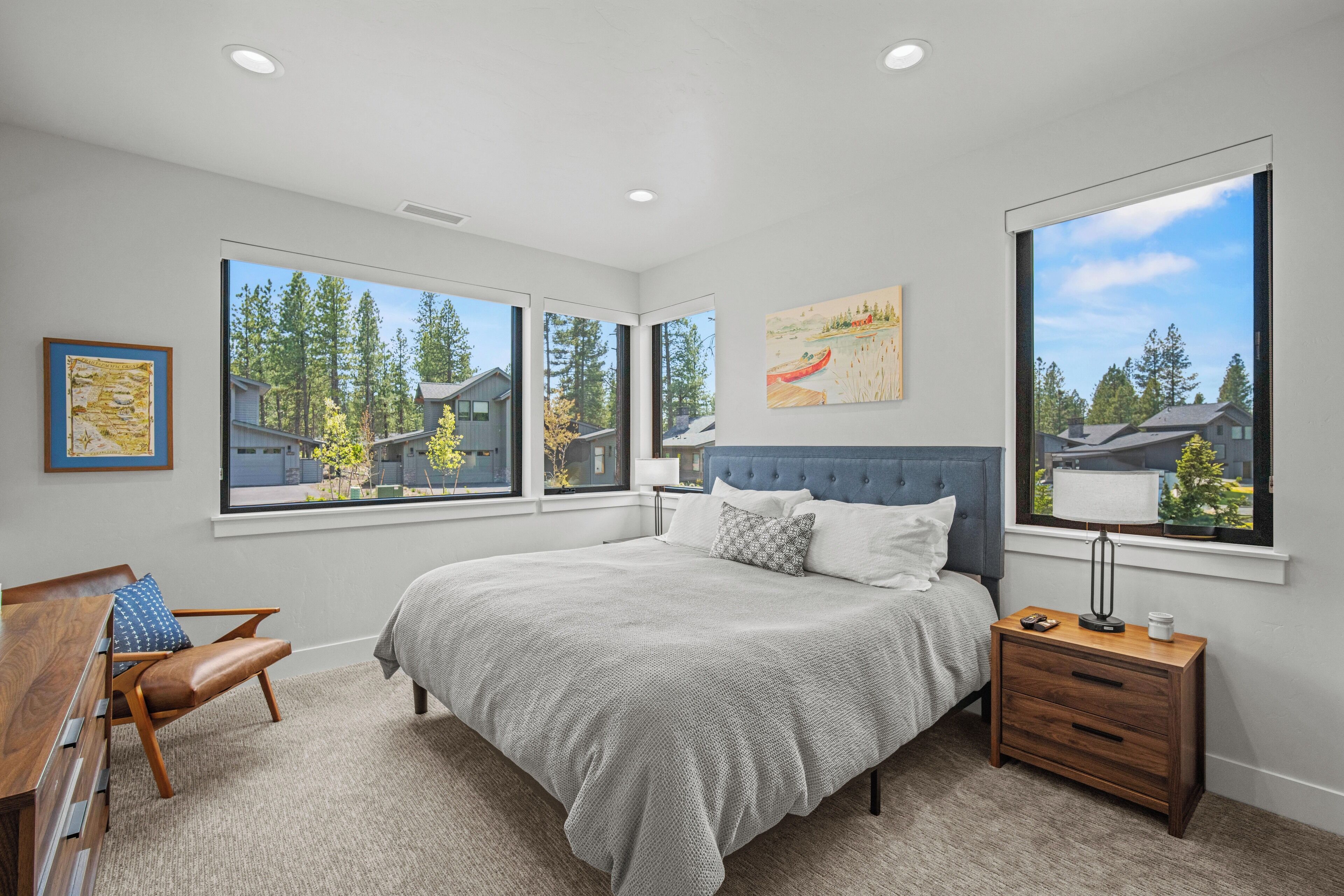 A bedroom with a large bed, a desk, and a chair. There are three windows providing views of houses and trees.