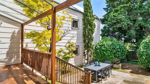 A covered outdoor patio with a wooden railing, a table and chairs, and a view of a house with a window and a tree.