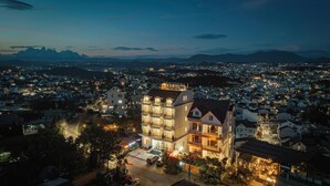 Un hôtel à plusieurs étages doté d’un balcon imposant au deuxième étage, situé dans un quartier urbain densément peuplé, entouré de lampadaires et avec une vue sur des montagnes lointaines.