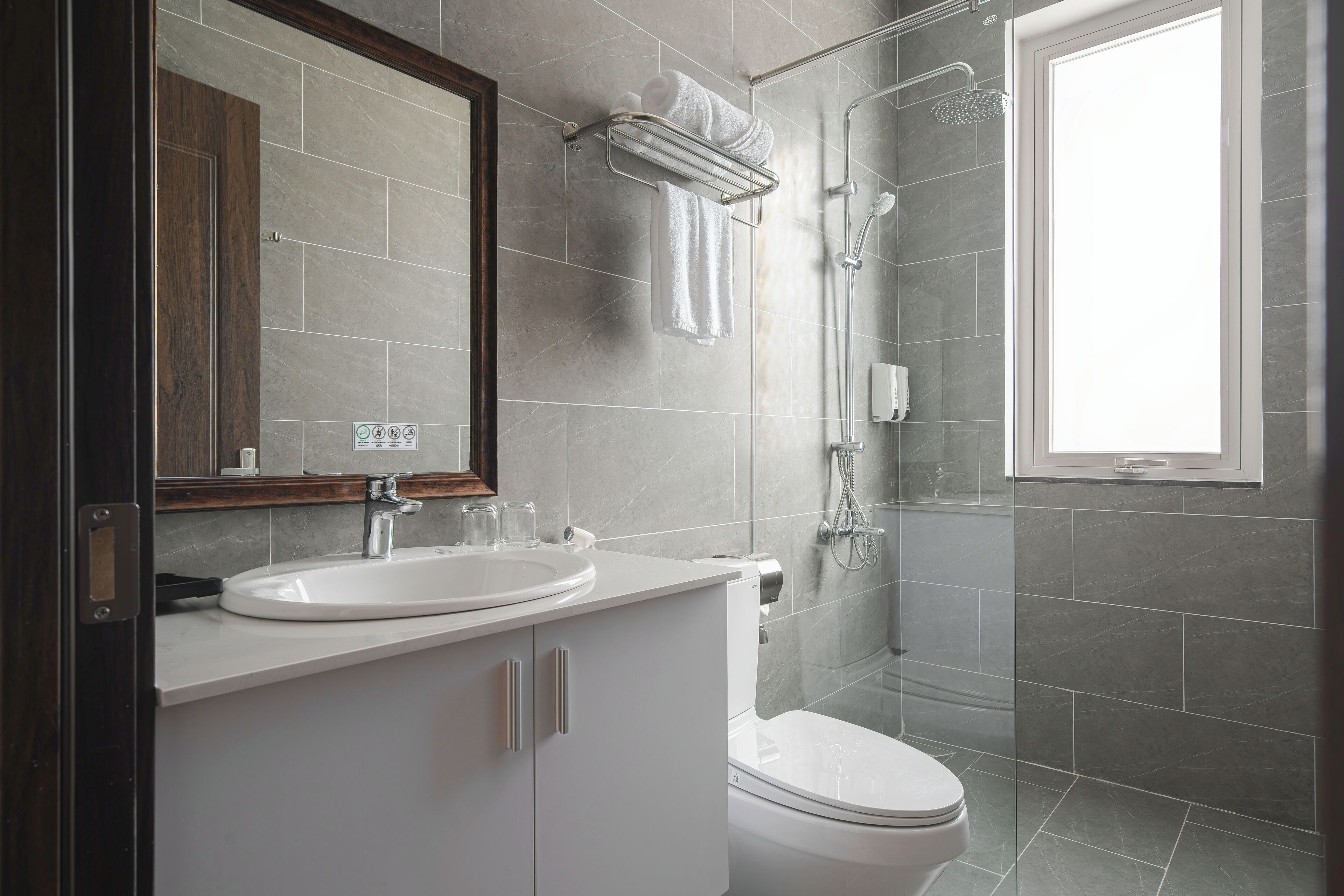 A modern bathroom with a white sink, mirror, and toilet, a glass shower enclosure, and a window providing natural light.