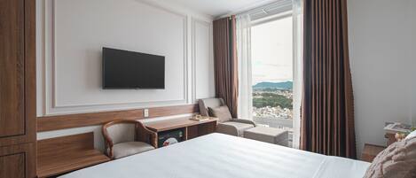 A hotel room with a large bed, a desk with a chair, a television mounted on the wall, and a window with curtains.