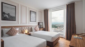 Une chambre d’hôtel avec deux lits, une tête de lit en bois, une table de chevet avec une lampe, une table en bois et une vue sur le paysage urbain à travers de grandes fenêtres.