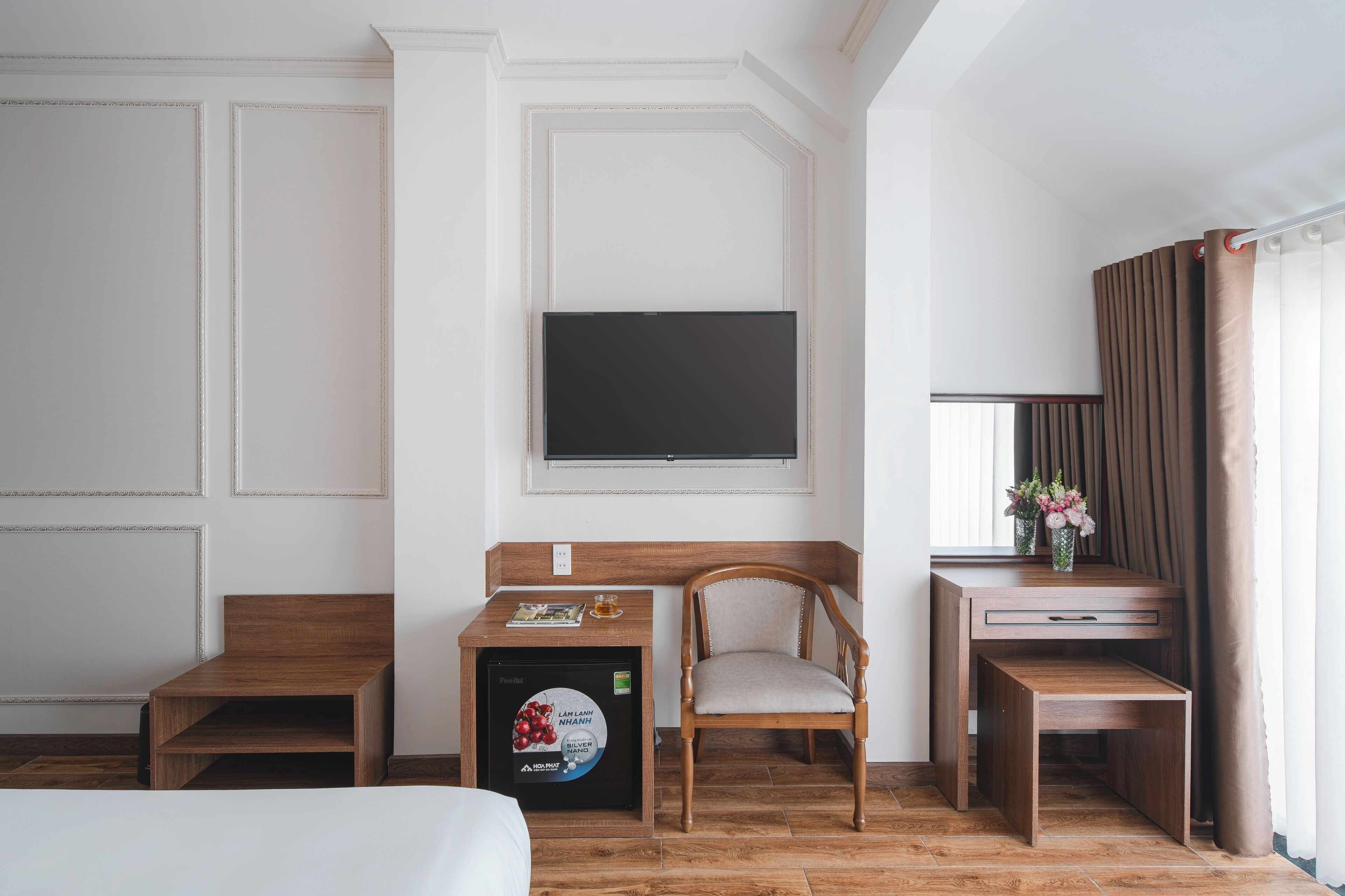 A hotel room with a bed, a wooden desk with a chair, a small refrigerator, a flat-screen TV mounted on the wall, and a window with curtains.