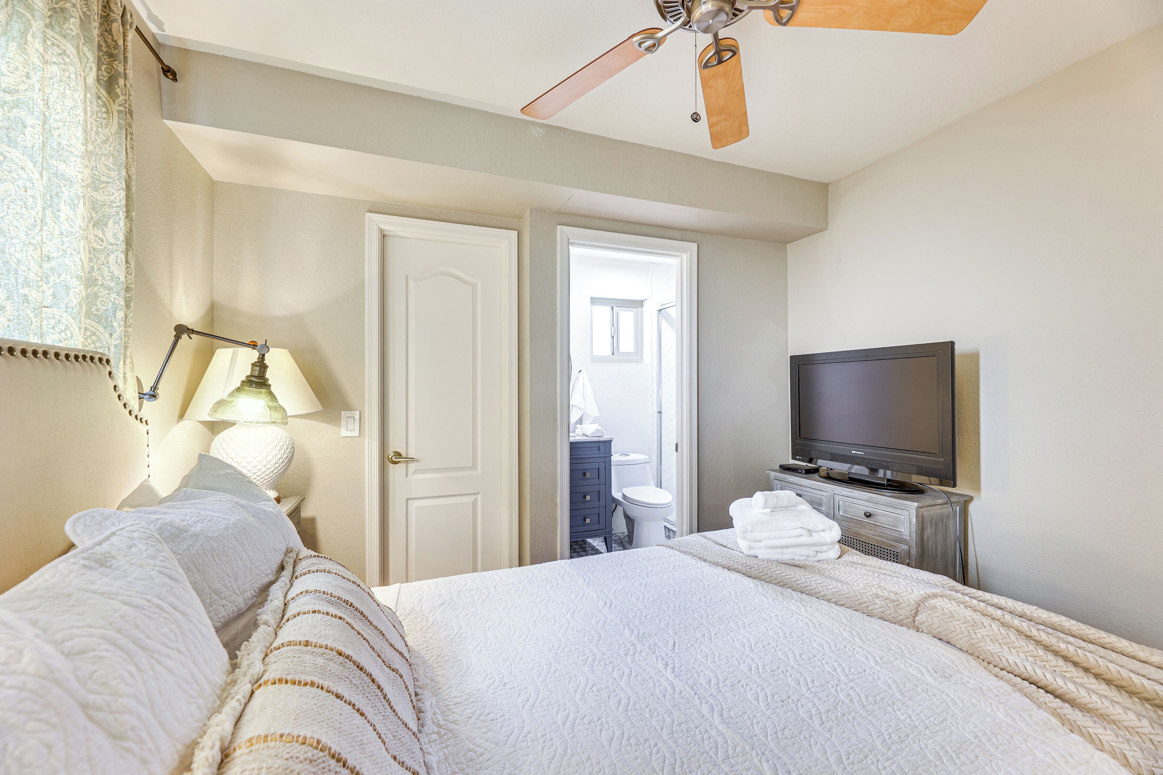 A bedroom with a bed, a television, a ceiling fan, and a bathroom visible through an open door.