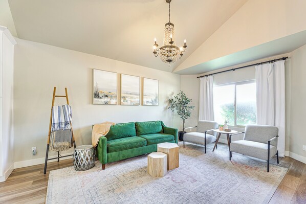 A spacious living room with a green sofa, a grey armchair, a wooden coffee table, and a chandelier. - Val Vista Lakes Group Getaway: Lake & Pool Access! (Gilbert)