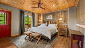 Ein Schlafzimmer mit Holzbalkendecke, einem Bett, einem Nachttisch, einer Bank und einem Fenster mit Vorhängen.