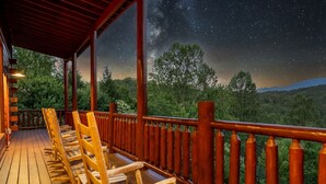 Eine Holzterrasse mit Stühlen und Geländer, mit Blick auf eine bewaldete Landschaft unter einem sternenübersäten Nachthimmel.