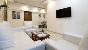 A modern living room with white sofas, a coffee table, a ceiling fan, and a mounted TV. - 3BHK Master Suite (Gagal Home) (Mumbai)