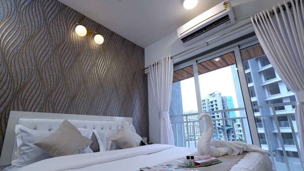A hotel room with a large bed, white bedding, a patterned wall, an air conditioning unit, and a view of buildings through the window. - 3BHK Master Suite (Gagal Home) (Mumbai)