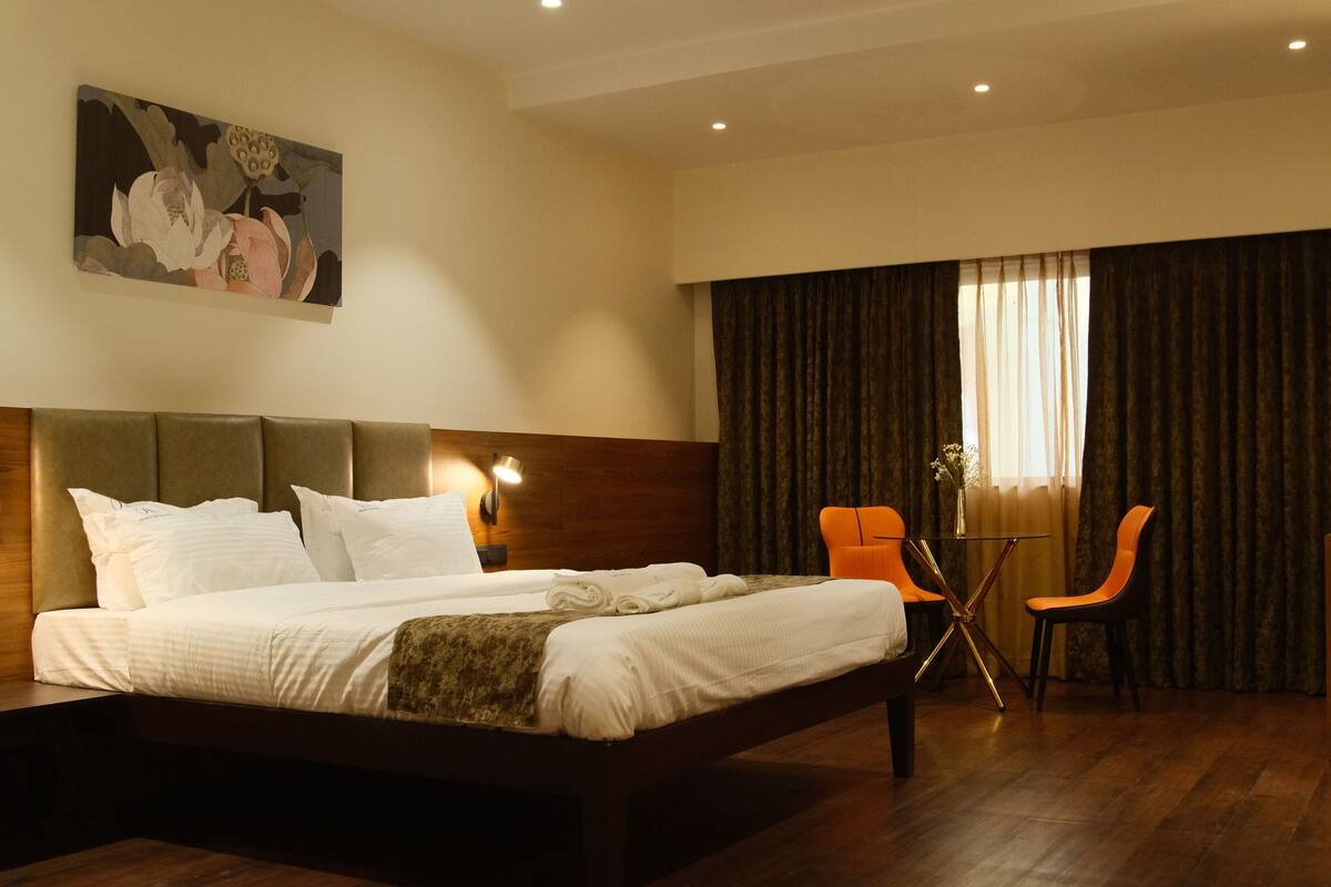 A hotel room with a large bed, a headboard, two chairs, a small table, and a painting on the wall.