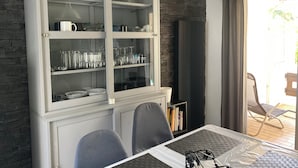 Ein Essbereich mit einem weißen Schrank, der Glaswaren und Geschirr beherbergt, einem Tisch mit Schutzdecke und Blick ins Freie durch eine Glastür.