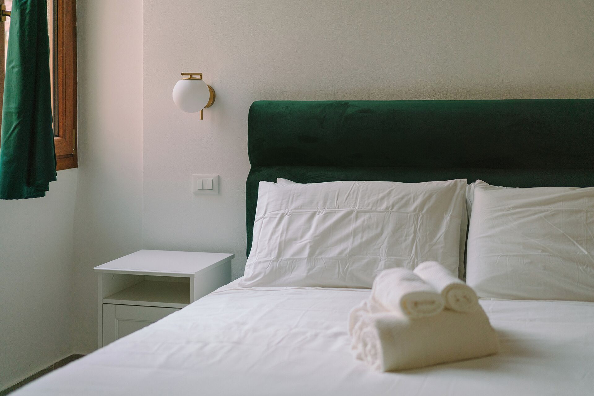 A neatly made bed with white linens and rolled towels, a green upholstered headboard, a white bedside table, and a wall-mounted light fixture.