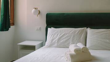 A neatly made bed with white linens and rolled towels, a green upholstered headboard, a white bedside table, and a wall-mounted light fixture.