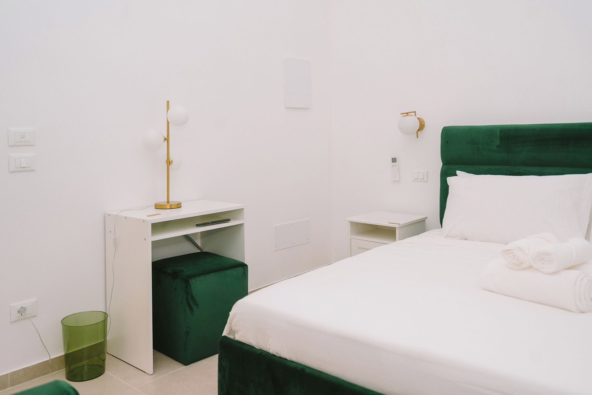 A minimalist hotel room with a green upholstered bed, a white desk with a lamp, and a green trash bin.