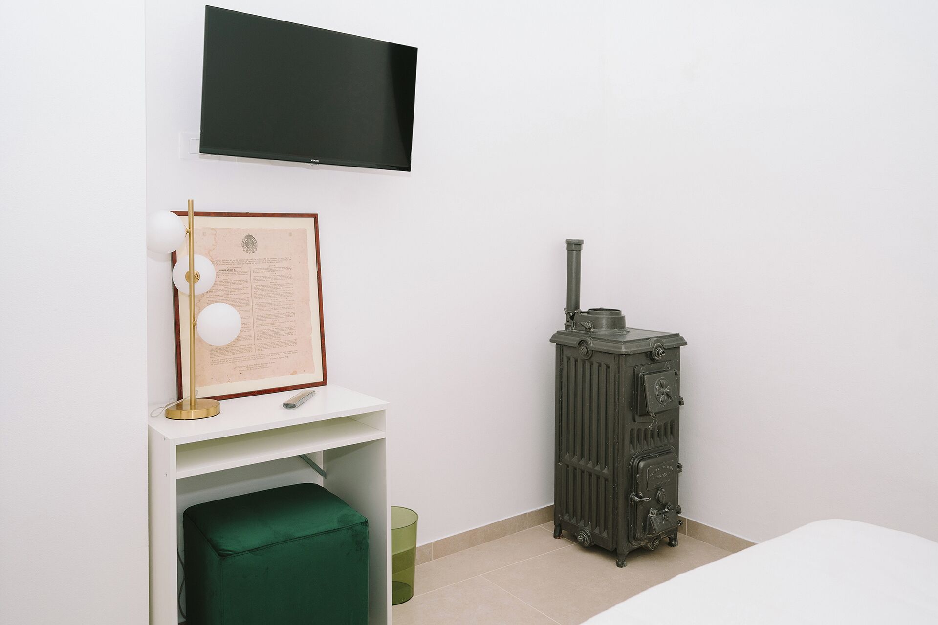 A room with a flat-screen TV mounted on the wall, a framed document on a shelf, a vintage-style radiator, and a green ottoman.