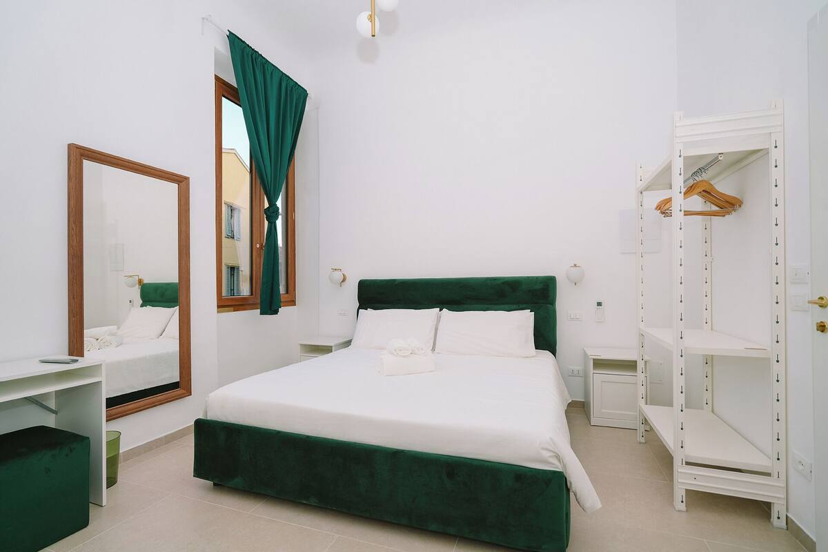 A neatly arranged bedroom with a green upholstered bed, a white bedside table, a mirror, a wardrobe, and a window with curtains.