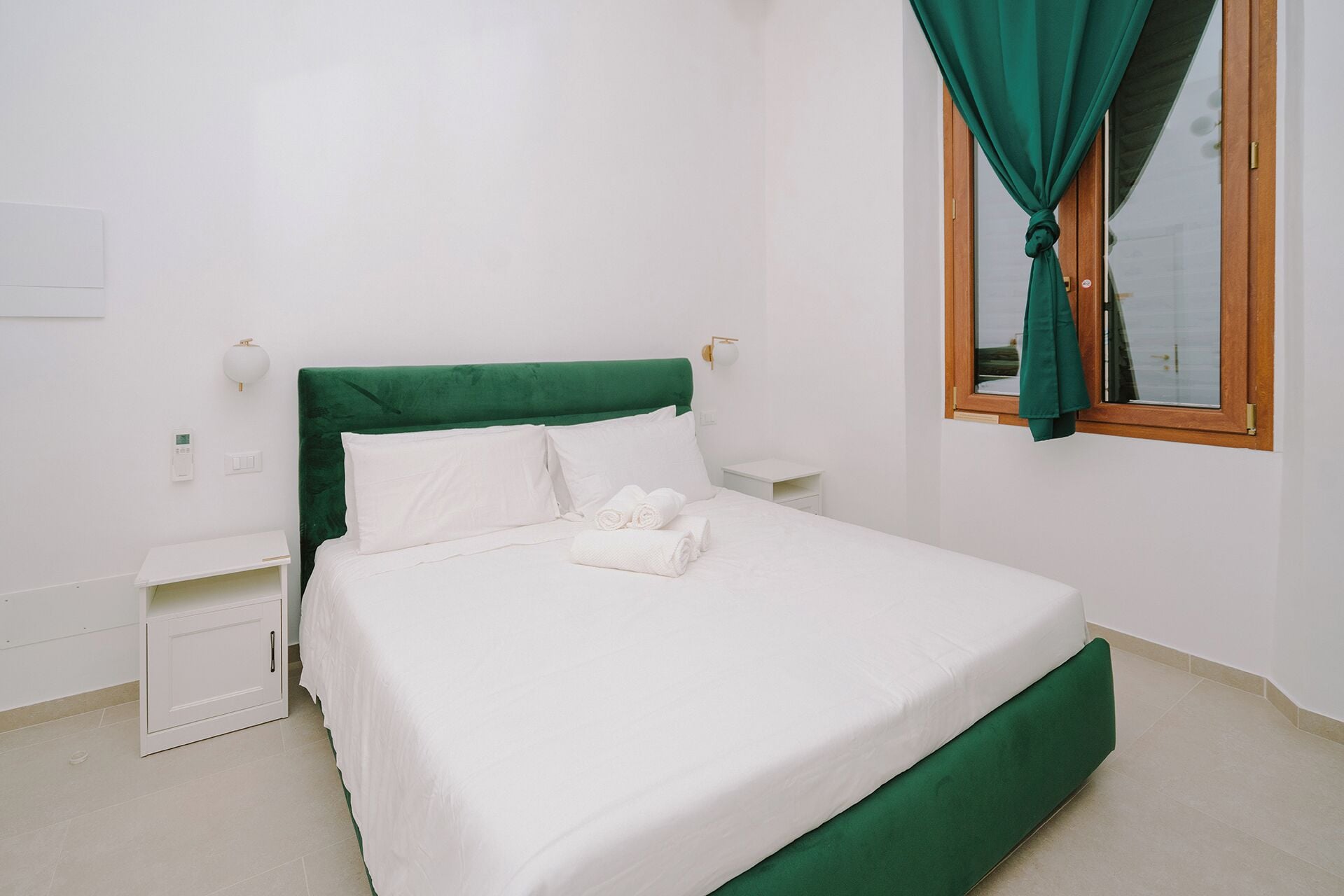 A neatly made bed with white linens and a green headboard in a simple, clean room with white walls and a window with green curtains.