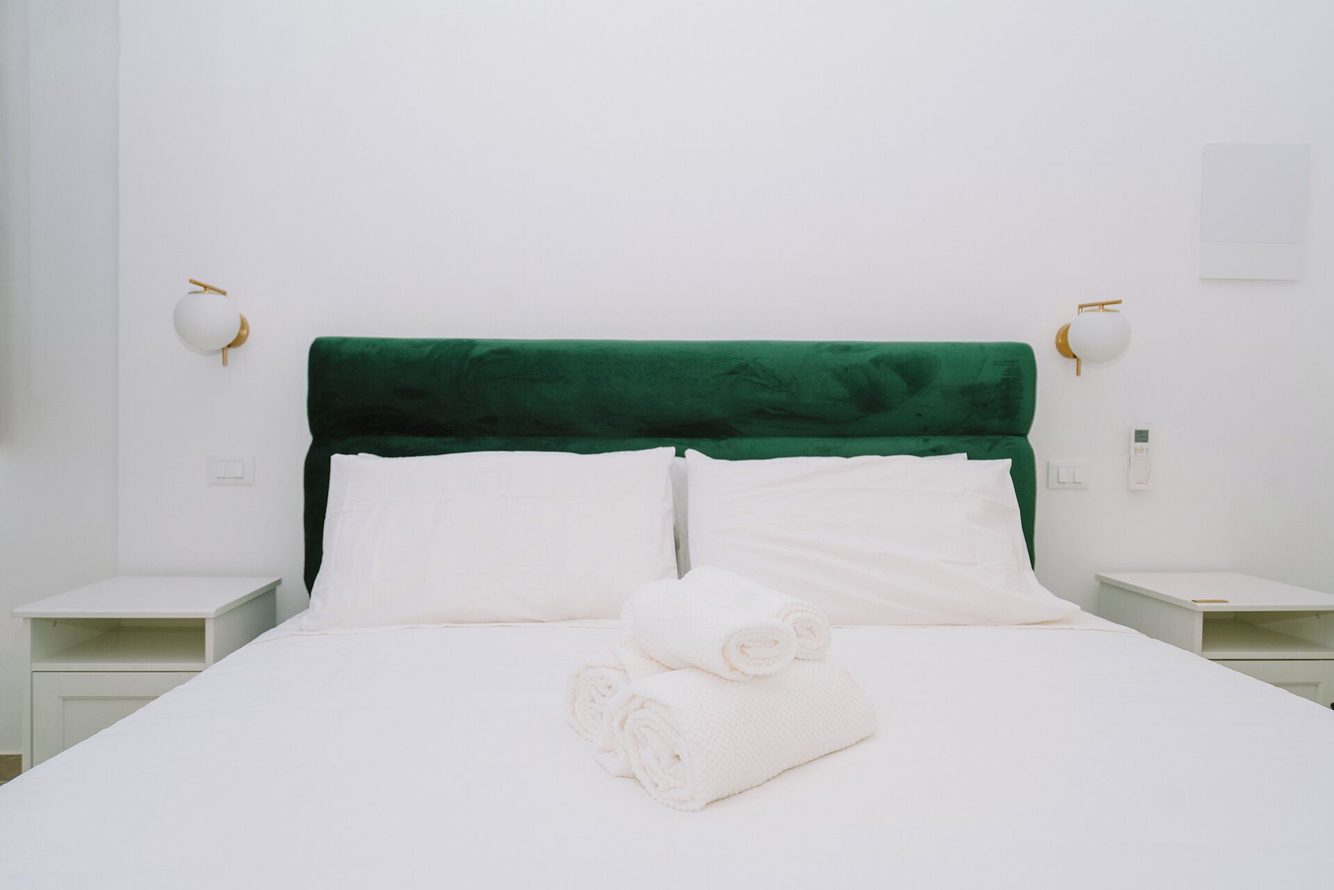 A bed with a green headboard, white pillows, and a rolled white towel. There are two bedside tables with lamps.