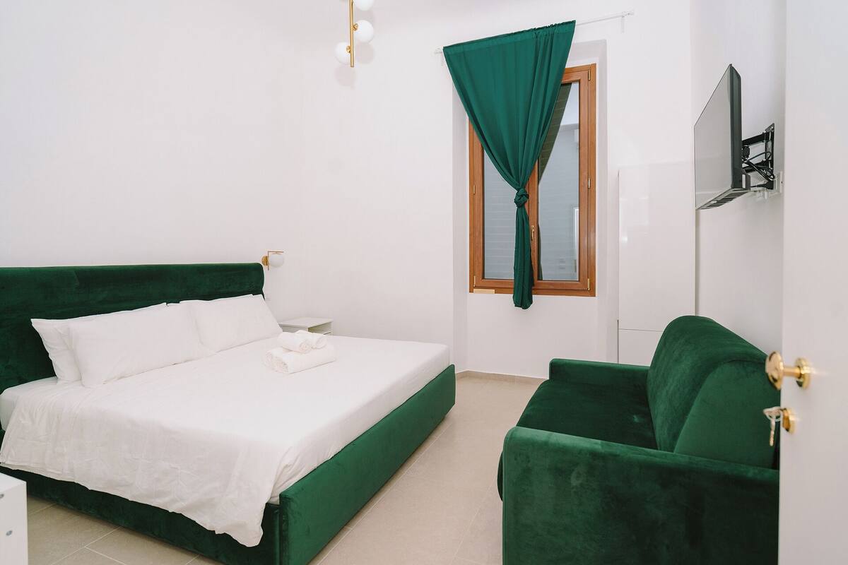 A room with a bed, a chair, a window with curtains, and a TV mounted on the wall.