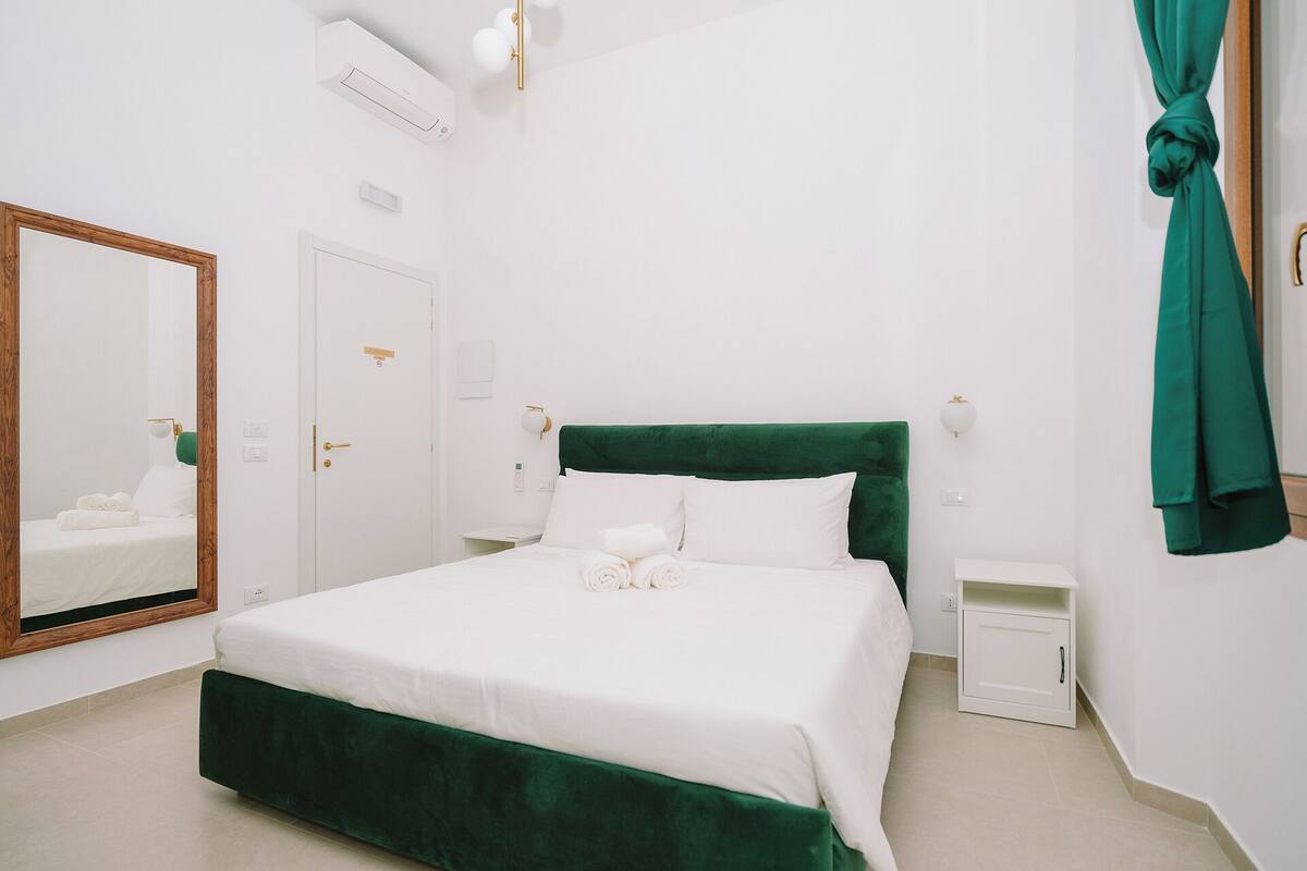 A neatly arranged hotel room with a large bed, a bedside table, a mirror, and a green curtain.