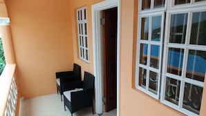 Un balcon avec deux fauteuils en osier noir, une table blanche et une fenêtre à cadre blanc donnant sur une voiture bleue.