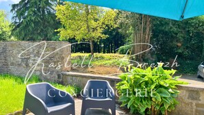 Un espace extérieur aménagé avec deux chaises et une table, un mur en pierre et un jardin où poussent différentes espèces de plantes.