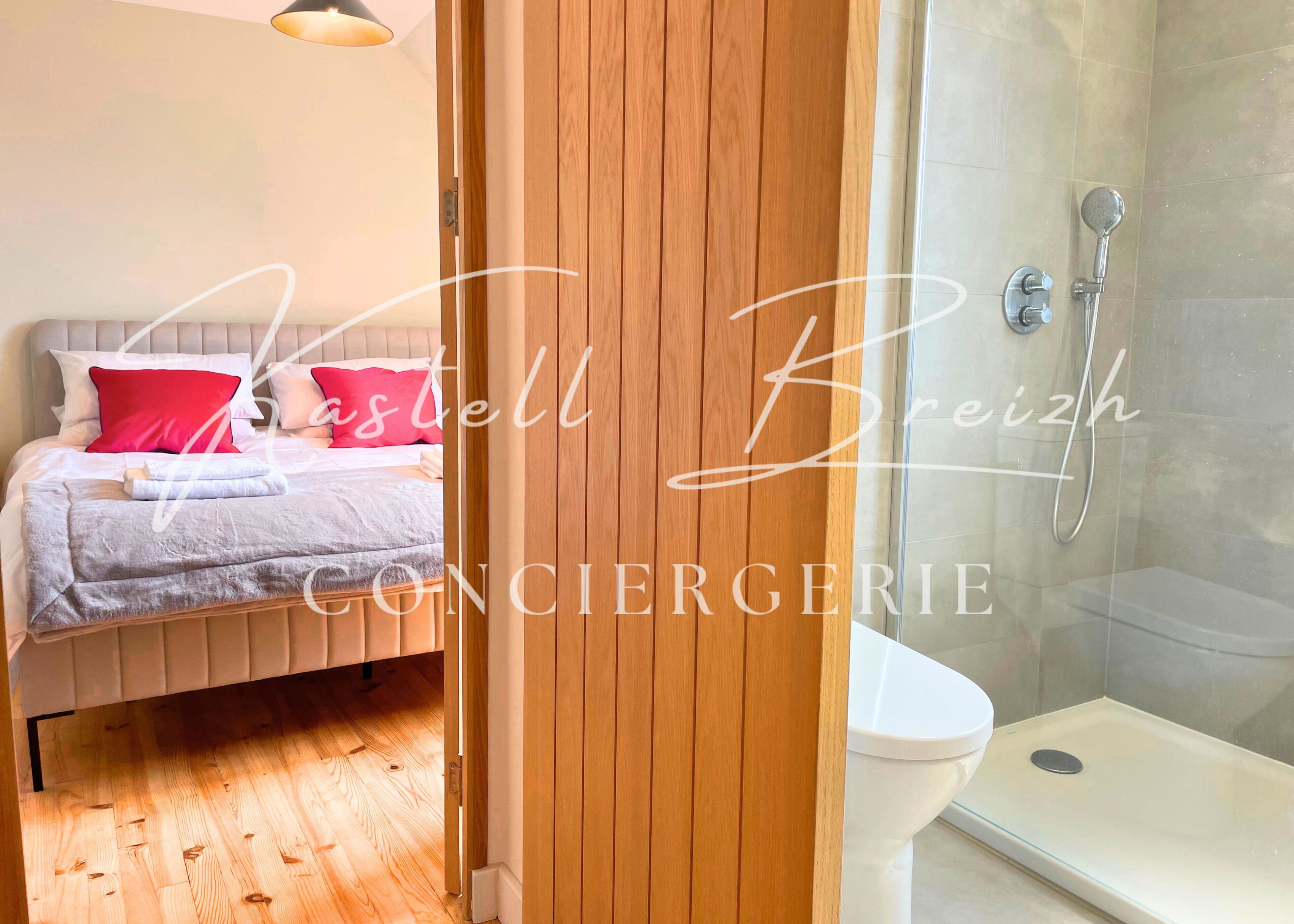 A bedroom with a bed, a bedside table, and a red pillow. There is a wooden door leading to a bathroom with a shower and a toilet.